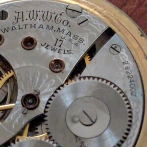 Puede incluir: Primer plano del mecanismo de un reloj de bolsillo antiguo. La esfera del reloj muestra el texto "A.W.W. Co. Waltham, Mass. U.S.A. 17 Jewels." Los intrincados engranajes y componentes son visibles, con un marco dorado.