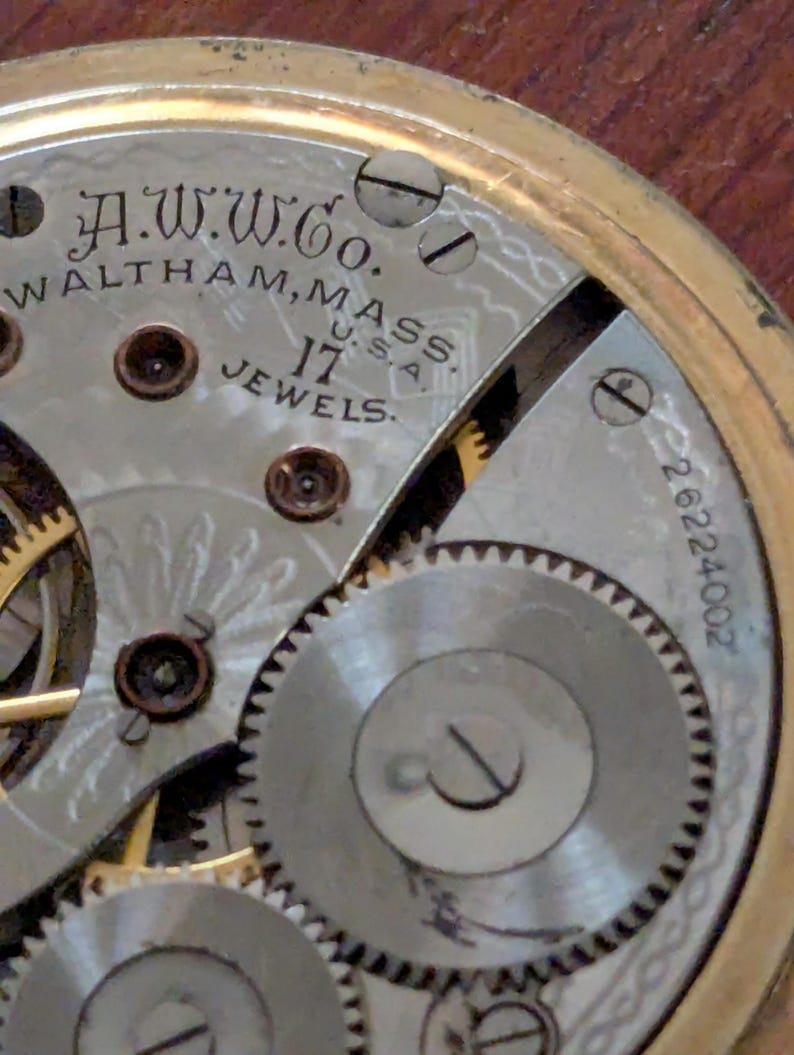 Puede incluir: Primer plano del mecanismo de un reloj de bolsillo antiguo. La esfera del reloj muestra el texto "A.W.W. Co. Waltham, Mass. U.S.A. 17 Jewels." Los intrincados engranajes y componentes son visibles, con un marco dorado.