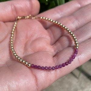 May include: A gold and purple beaded bracelet with a clasp fastening. The bracelet is made with small, round beads.