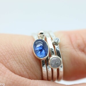 K&ouml;nnte beinhalten: Ein Satz von vier Silberringen mit verschiedenen Steinen. Der oberste Ring hat einen blauen ovalen Stein, der zweite Ring einen kleinen, klaren, runden Stein und der dritte Ring einen wei&szlig;en ovalen Stein. Der unterste Ring ist aus einfachem Silber.