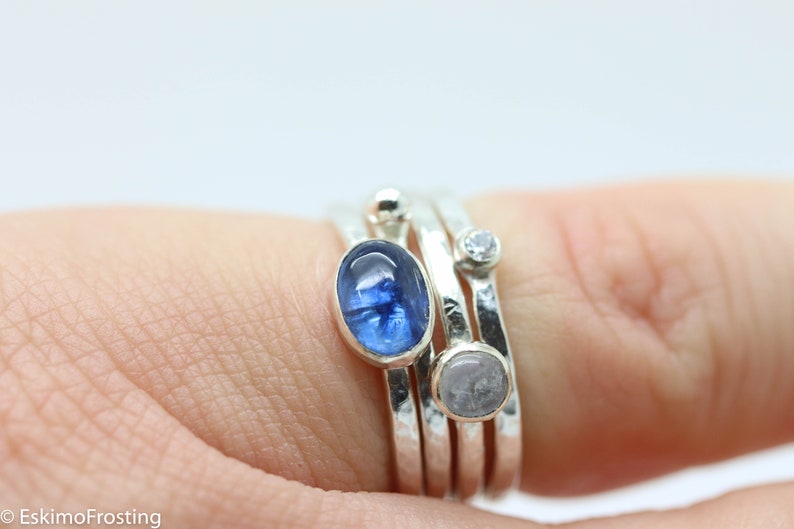 K&ouml;nnte beinhalten: Ein Satz von vier Silberringen mit verschiedenen Steinen. Der oberste Ring hat einen blauen ovalen Stein, der zweite Ring einen kleinen, klaren, runden Stein und der dritte Ring einen wei&szlig;en ovalen Stein. Der unterste Ring ist aus einfachem Silber.