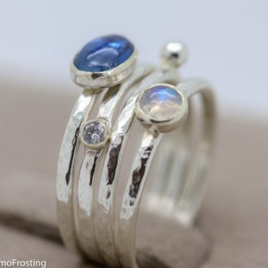 K&ouml;nnte beinhalten: Ein Satz von vier Silberringen mit geh&auml;mmerten Texturen. Die Ringe haben drei Edelsteine: ein blaues Oval, ein wei&szlig;er Rundstein und ein wei&szlig;es Oval mit irisierendem Schimmer.