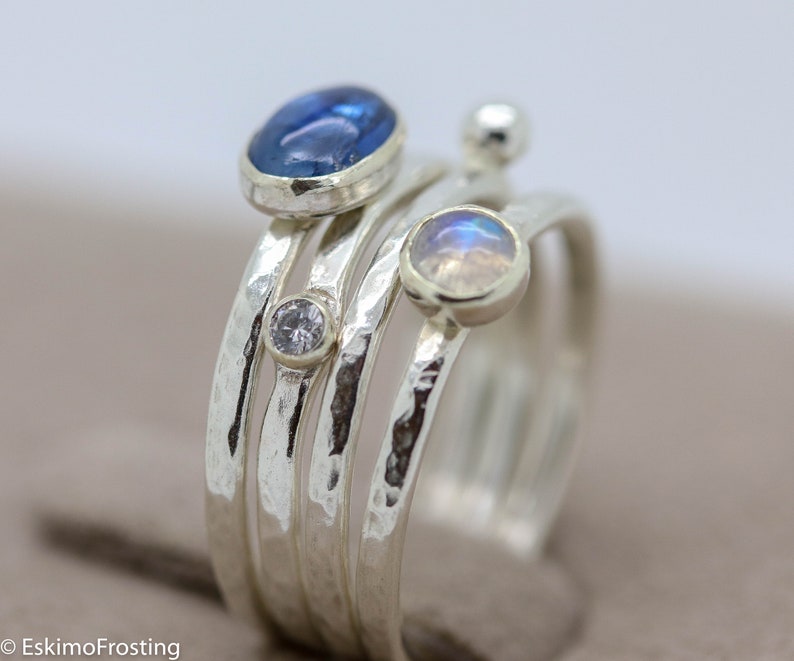 K&ouml;nnte beinhalten: Ein Satz von vier Silberringen mit geh&auml;mmerten Texturen. Die Ringe haben drei Edelsteine: ein blaues Oval, ein wei&szlig;er Rundstein und ein wei&szlig;es Oval mit irisierendem Schimmer.