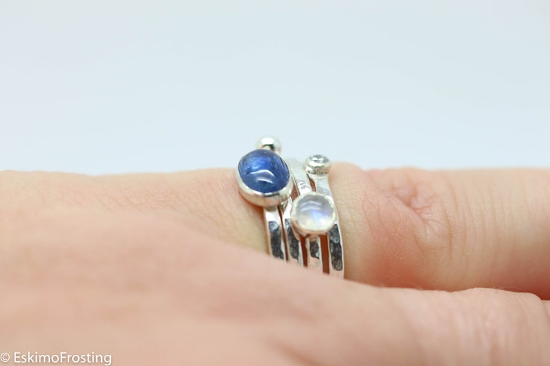 K&ouml;nnte beinhalten: Ein Satz aus drei Silberringen mit Edelsteinen. Der obere Ring hat einen blauen ovalen Edelstein, der mittlere Ring einen klaren runden Edelstein und der untere Ring einen wei&szlig;en ovalen Edelstein.