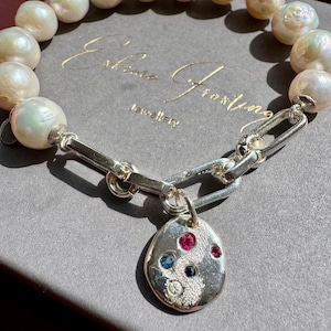 May include: A pearl bracelet with a silver chain and a pendant. The bracelet features large, irregularly shaped pearls. The silver pendant is oval and adorned with colourful gemstones, including red, blue, and clear stones. The bracelet is displayed on a grey surface.