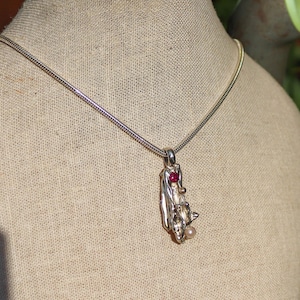May include: A silver chain necklace with a pendant featuring a textured silver and gold design with a red gemstone and a pearl.