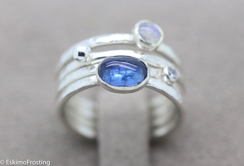K&ouml;nnte beinhalten: Ein silberner Ring mit vier B&auml;ndern, besetzt mit einem blauen Edelstein und zwei kleineren wei&szlig;en Edelsteinen. Der Ring hat eine geh&auml;mmerte Textur.
