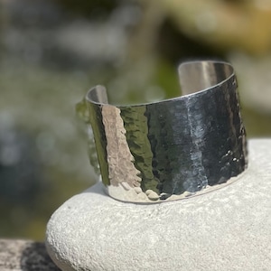 Könnte beinhalten: Ein silbernes, gehämmertes Armreif-Armband. Das Armband ist breit und hat eine strukturierte Oberfläche.