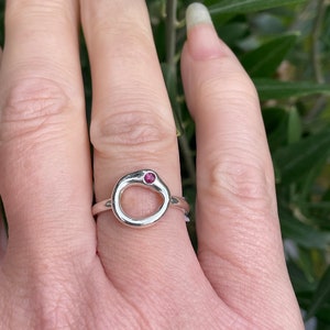 May include: A silver ring with a small, round, pink gemstone set in the centre of an open circle.