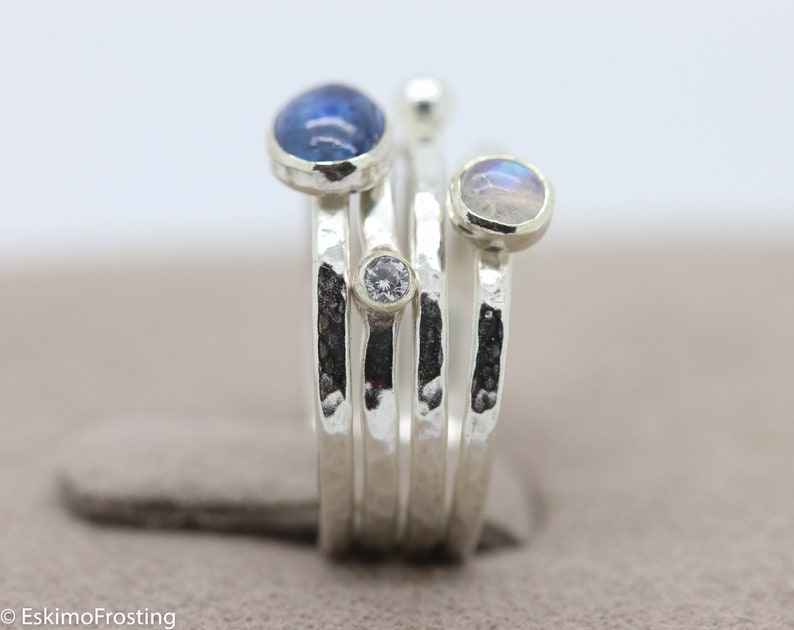 K&ouml;nnte beinhalten: Ein Silberring mit vier Steinen. Der Ring besteht aus mehreren B&auml;ndern mit einer geh&auml;mmerten Textur. Die Steine sind ein blauer Edelstein, ein wei&szlig;er Edelstein und zwei schillernde Mondsteine.
