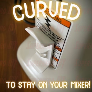 May include: White plastic 3D printed stand with a curved design. The stand is designed to hold a kitchen mixer in place. The stand is attached to a white surface. The text "CURUED" is in yellow letters on a white background. The text "TO STAY ON YOUR MIXER!" is in yellow letters on a white background. The text on the yellow warning label reads "Electrical Shock Hazard. Unplug before servicing. Failure to do so can result in shock."