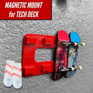 May include: A red rectangular magnetic mount for Tech Deck fingerboards featuring four raised rectangular tabs. Two fingerboards are attached to the mount. Two white adhesive strips are positioned to the left of the mount.