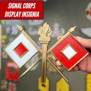 May include: A gold-colored Signal Corps display insignia featuring a central torch flanked by two crossed flags. The left flag is white with a red square center, and the right flag is red with a white square center. The text reads SIGNAL CORPS DISPLAY INSIGNIA.