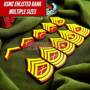 May include: A collection of USMC enlisted rank insignia in yellow and red, with the text "USMC ENLISTED RANK MULTIPLE SIZES" in a red box. The insignia are arranged on a green fabric.