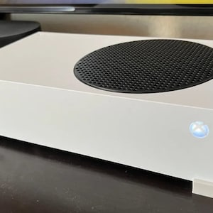 May include: A white Xbox Series S console with a black circular vent on top. The console has a glowing Xbox logo and is elevated on white feet. The design is minimalist and modern.