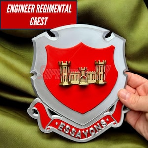 May include: A silver, red, and gold Engineer Regimental Crest. The crest features a red shield with a gold castle emblem. The text "ENGINEER REGIMENTAL CREST" is displayed at the top. The word "ESSAYONS" is displayed at the bottom.