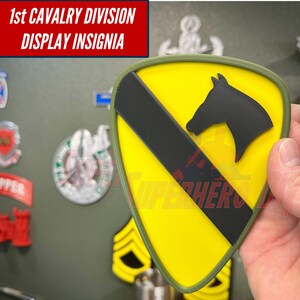 May include: A shield-shaped 1st Cavalry Division display insignia featuring a bright yellow background, a diagonal black stripe, and a black horse head silhouette. The emblem is bordered by an olive green edge and includes the text 1st CAVALRY DIVISION DISPLAY INSIGNIA.