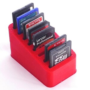 May include: A red 3D-printed SD card holder with multiple slots, holding various black SD cards with brand labels like SanDisk and Lexar. The holder is rectangular and designed to organize memory cards.