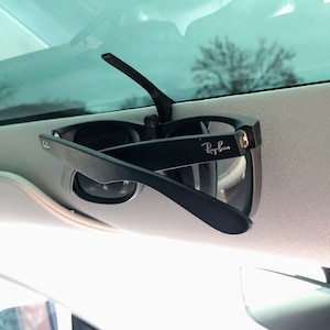 May include: Black Ray-Ban sunglasses clipped to a car visor. The sunglasses are in a classic style with dark lenses and the Ray-Ban logo visible on the temple. The visor is light gray.