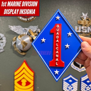 May include: A diamond-shaped 1st Marine Division display insignia featuring a blue background with a large red number one. The word GUADALCANAL is printed vertically in white on the red number. Five white stars are arranged around the center.