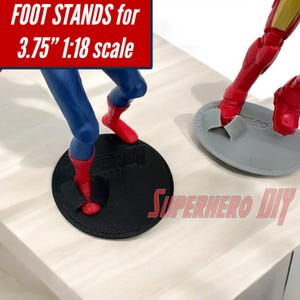 May include: Black and gray foot stands for 3.75-inch 1:18 scale action figures. The stands are round and designed to support the figures. The text "FOOT STANDS for 3.75" 1:18 scale" is in a red box. The text "SUPERHERO DIY" is in red.