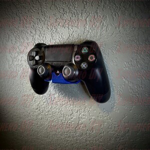 May include: A black PlayStation 4 controller with blue accents is mounted on a wall. The controller has a textured surface and a variety of buttons, including the PlayStation logo, the directional pad, the action buttons, and the share button.