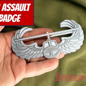May include: A silver Air Assault Badge with wings and a helicopter in the center. The badge is held in a hand against a green fabric background. The text "AIR ASSAULT BADGE" is in red letters on a white background.