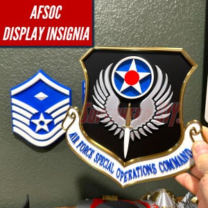 May include: A black, gold, and white Air Force Special Operations Command display insignia featuring a central silver winged emblem with a dagger and a blue star with a red circle. A blue and white chevron insignia is visible in the background.