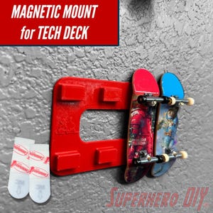 May include: Red magnetic mount for Tech Deck skateboards, holding two miniature skateboards with red and blue decks. The mount is attached to a gray wall with Command strips. Text on the mount reads "MAGNETIC MOUNT for TECH DECK".