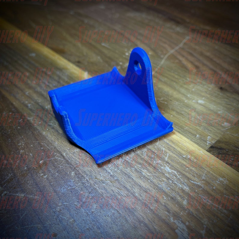 May include: A blue 3D printed plastic wall mount with a hole at the top. The mount is designed to hold a small object, such as a camera or a phone.