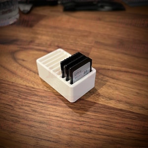 May include: White plastic storage case for memory cards. The case has four slots and is holding three memory cards. One of the cards is labeled "Ultra 60MB/s".