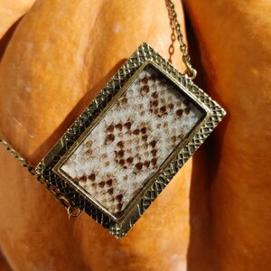 May include: A gold-toned necklace with a rectangular pendant featuring a snakeskin pattern. The pendant is framed with a decorative border.