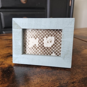 May include: A small, rectangular, light blue wooden picture frame. The frame surrounds a patterned, off-white and brown material with a white heart shape in the center. The frame sits on a dark wooden surface.