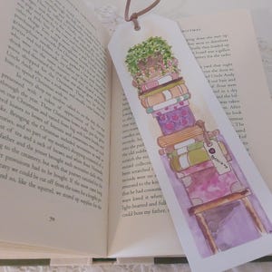 May include: A watercolor bookmark featuring a stack of books with a potted plant on top. The bookmark is on a white background and has the word "Books" written on it.