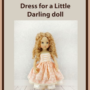 Puede incluir: Patrón PDF y foto tutorial para un vestido para una pequeña muñeca querida. La muñeca lleva un vestido de color melocotón con ribete de encaje blanco y zapatos blancos. La muñeca tiene el pelo rubio rizado.