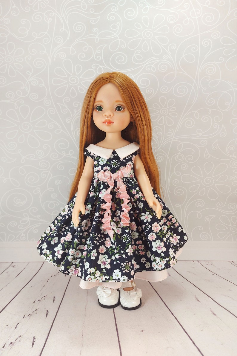 May include: A doll with long red hair wearing a blue and white floral dress with a pink bow. The doll is wearing white shoes with a pink bow.