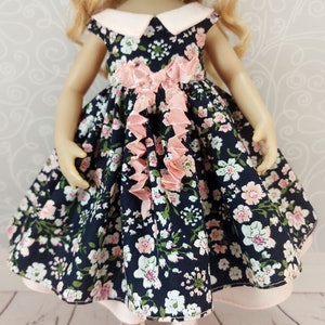 May include: A doll wearing a navy blue dress with a pink floral print and a pink bow. The dress has a white collar and a pink ruffle trim. The doll is wearing white shoes.
