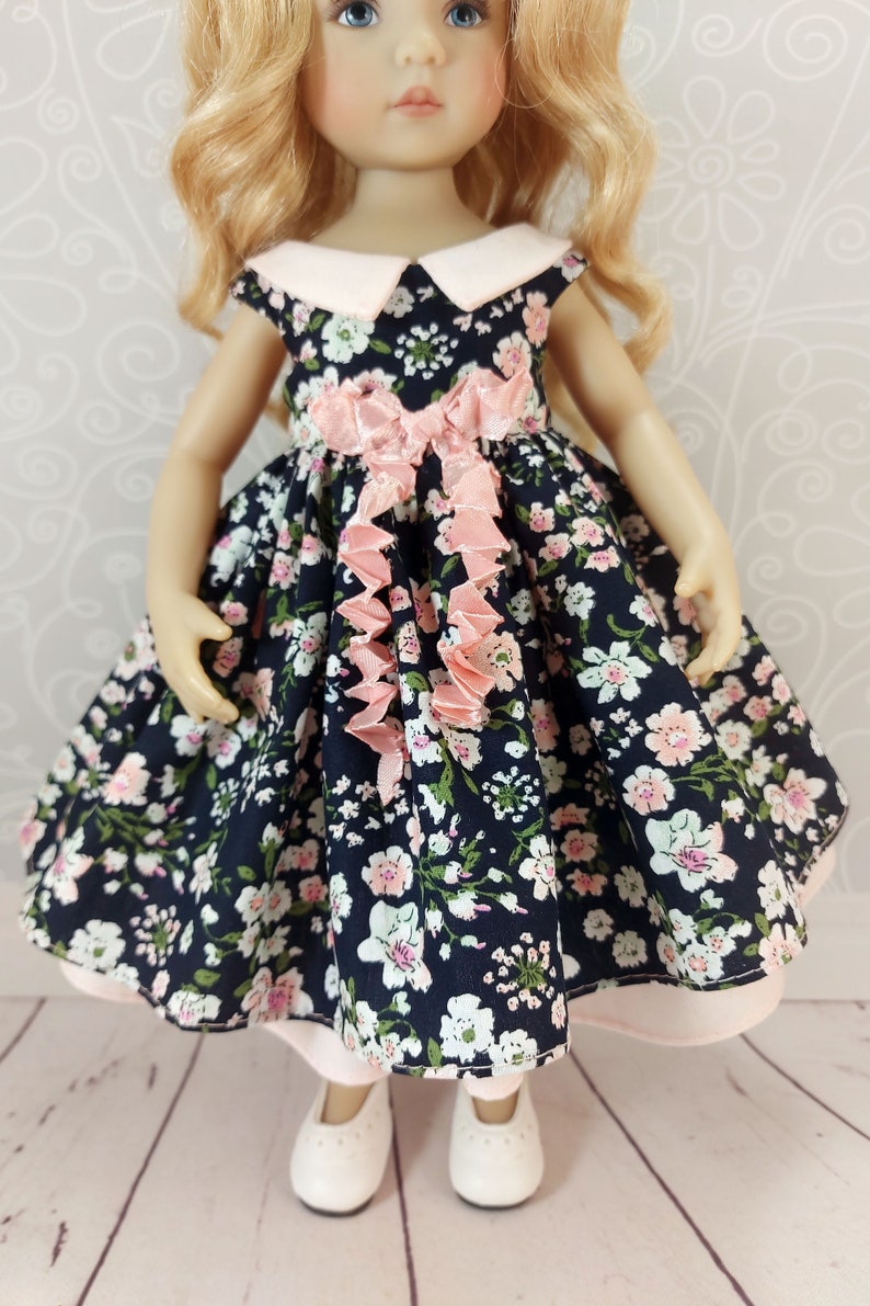 May include: A doll wearing a navy blue dress with a pink floral print and a pink bow. The dress has a white collar and a pink ruffle trim. The doll is wearing white shoes.