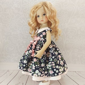 May include: A doll wearing a navy blue dress with a pink floral pattern and a white collar. The dress has a pink ribbon tied at the waist. The doll has blonde curly hair and blue eyes. The doll is standing on a white wooden floor.