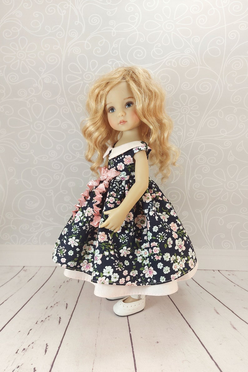 May include: A doll wearing a navy blue dress with a pink floral pattern and a white collar. The dress has a pink ribbon tied at the waist. The doll has blonde curly hair and blue eyes. The doll is standing on a white wooden floor.