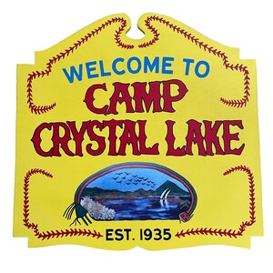 Puede incluir: Se&ntilde;al de madera amarilla con letras rojas y azules que dice "Bienvenido al Campamento Crystal Lake Est. 1935". La se&ntilde;al tiene un borde de puntadas de b&eacute;isbol rojas y blancas y una escena pintada de un lago con p&aacute;jaros volando por encima.