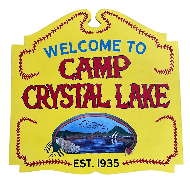 Puede incluir: Se&ntilde;al de madera amarilla con letras rojas y azules que dice "Bienvenido al Campamento Crystal Lake Est. 1935". La se&ntilde;al tiene un borde de puntadas de b&eacute;isbol rojas y blancas y una escena pintada de un lago con p&aacute;jaros volando por encima.