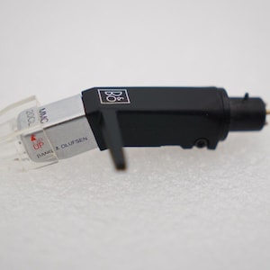 May include: A black and silver Bang & Olufsen MMC 20CL cartridge for a record player. The cartridge has a clear plastic stylus guard and the words "UP" and "BANG & OLUFSEN" are visible.