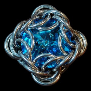 May include: A square, bright blue gemstone is the focal point of this jewellery piece. The gemstone is encased in a silver-coloured, chainmail-style setting. The setting is composed of interlocking, rounded metal links, creating a textured, geometric design. The background is black.