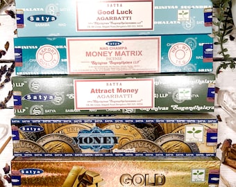 May include: Five boxes of Satya incense sticks. The boxes are stacked on top of each other. The top box is gold with the word "GOLD" in large letters. The second box is blue and green with the word "MONEY" in large letters. The third box is green with the words "Attract Money" in large letters. The fourth box is pink and blue with the words "NAG CHAMPA MONEY MATRIX" in large letters. The bottom box is blue with the words "Good Luck AGARBATTI" in large letters.