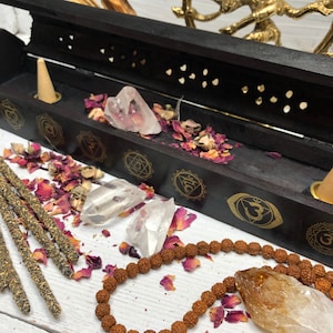 May include: Black wooden incense burner with gold chakra symbols and a small cone of incense. The burner is surrounded by rose petals, clear quartz crystals, and a brown mala.