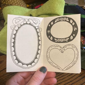 Puede incluir: Un peque&ntilde;o folleto abierto que muestra dibujos lineales en blanco y negro de marcos decorativos. Uno es ovalado con un lazo, otro redondo con detalles florales, y el tercero tiene forma de coraz&oacute;n formado por dos serpientes.