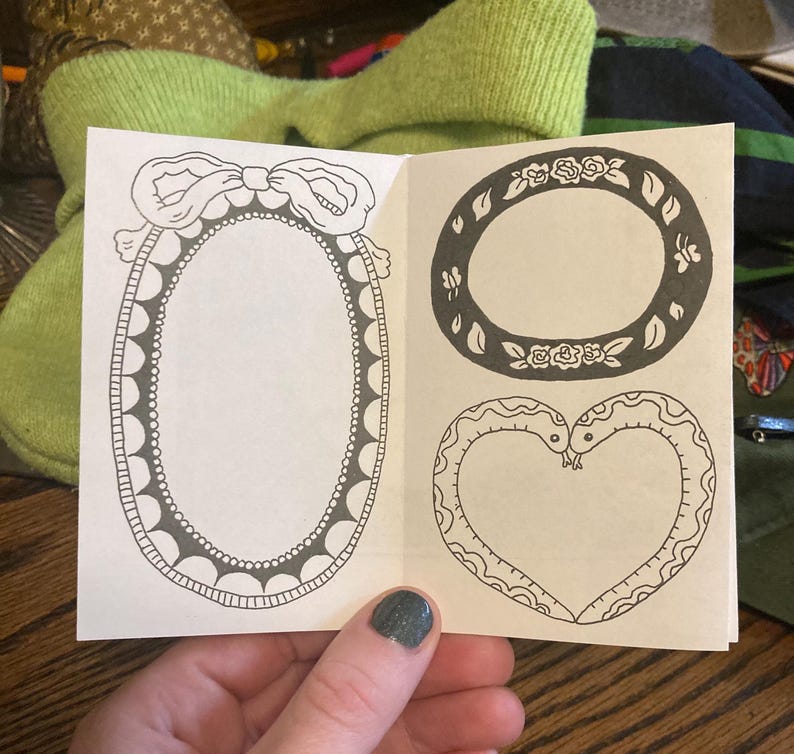 Puede incluir: Un peque&ntilde;o folleto abierto que muestra dibujos lineales en blanco y negro de marcos decorativos. Uno es ovalado con un lazo, otro redondo con detalles florales, y el tercero tiene forma de coraz&oacute;n formado por dos serpientes.