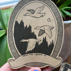 Peut inclure: Un autocollant avec un motif sur le thème de la nature. Il présente trois oies en vol au-dessus d'une silhouette de forêt, avec un croissant de lune dans le ciel. Le texte "FROM THE LIBRARY OF" est imprimé sur une bannière en bas. L'autocollant a un fond marron.