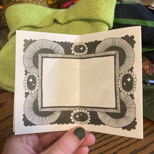 Puede incluir: Una tarjeta blanca plegada con un borde decorativo en blanco y negro. El borde presenta un patr&oacute;n repetitivo de l&iacute;neas curvas y formas ovaladas. La tarjeta se sostiene en una mano con esmalte de u&ntilde;as verde oscuro.